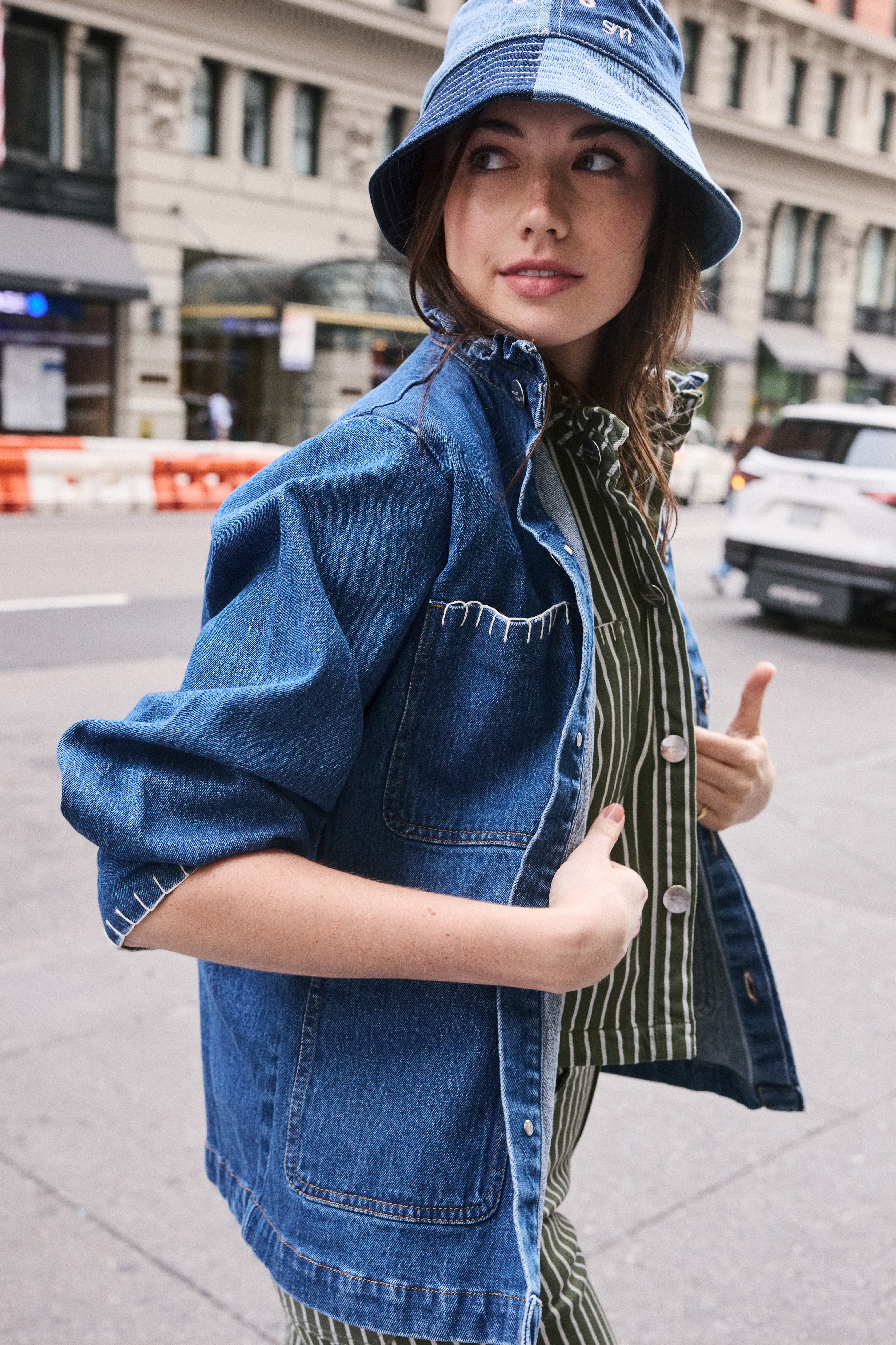woman wearing a blue denim jacket with a frill collar, white stitch detailing, button-up front, and four large pockets with a green and white striped denim shirt and jeans, and a denim bucket hat on a new york city street