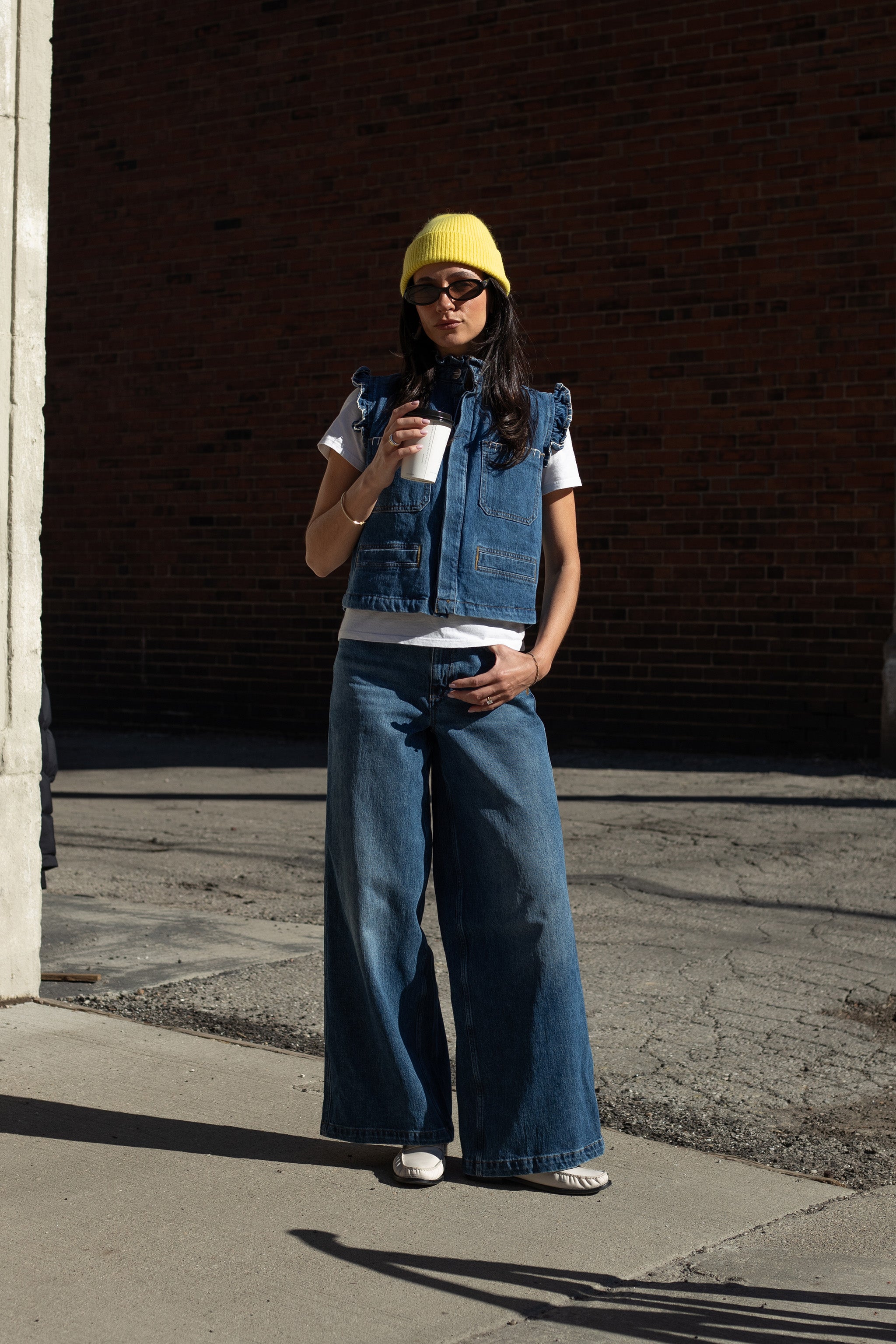woman wearing a blue denim vest/ waistcoat with our signature white stitch detailing, two large patch pockets, and a frill collar with blue jeans against a brick background