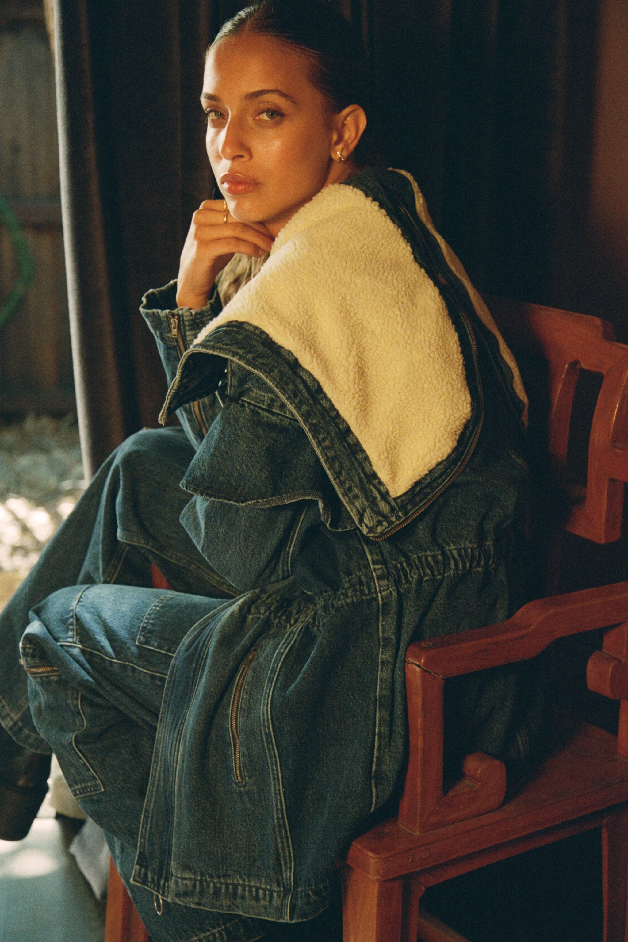 Close up of woman wearing a dark blue denim jacket with a white sherpa lined removable hood, waist cinch, and zip pocket details with dark blue jeans with cargo zip pockets, photographed from the back