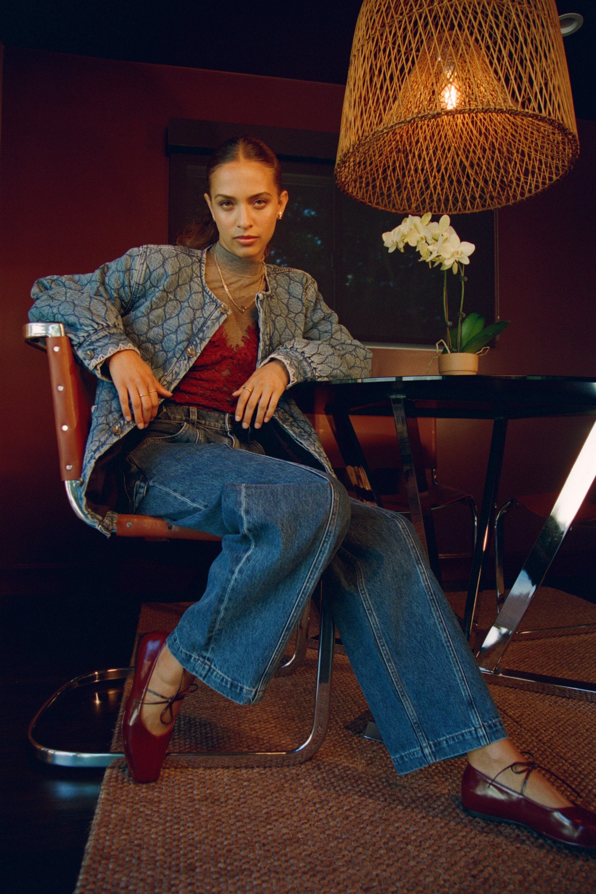 Woman sitting on chair in a quilted blue denim jacket with large pockets, a red shirt, blue barrel jeans, and red shoes