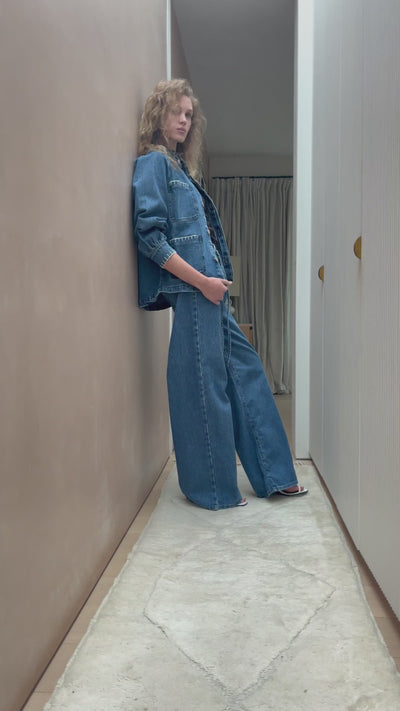 woman wearing a blue denim jacket with a frill collar, white stitch detailing, button-up front, and four large pockets with blue jeans