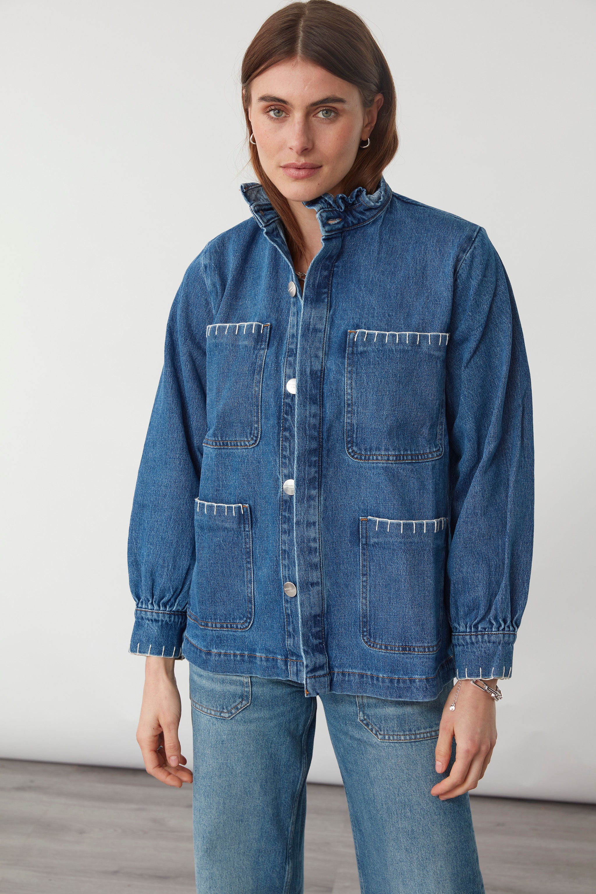 woman wearing a blue denim jacket with a frill collar and white blanket stitching on four large patch pockets and blue jeans against a plain background
