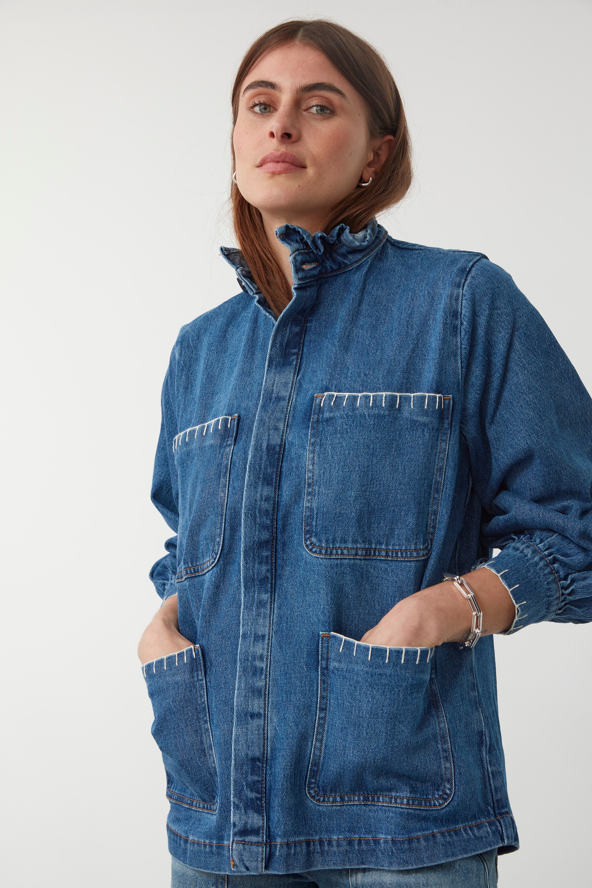 woman wearing a blue denim jacket with a frill collar and white blanket stitching on four large patch pockets and blue jeans against a plain background