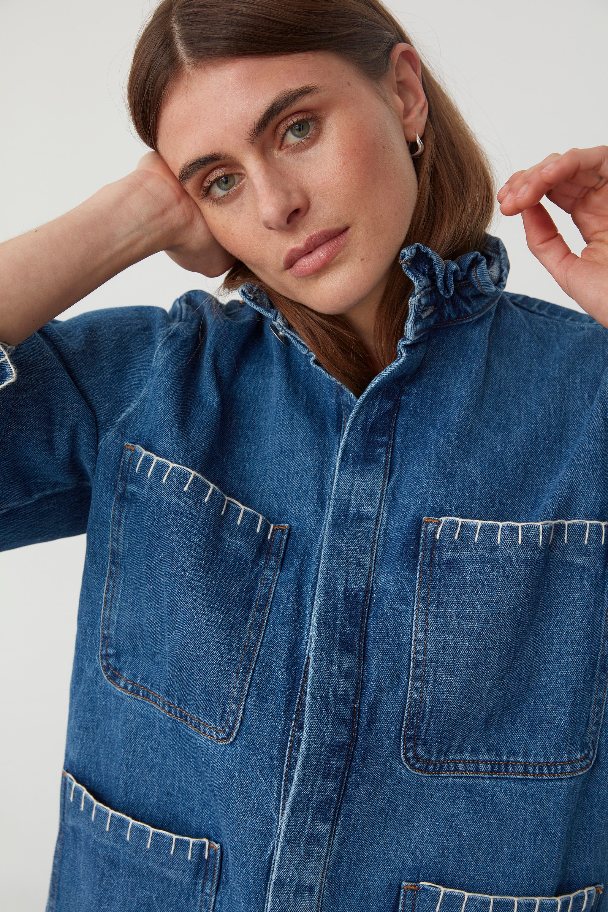 woman wearing a blue denim jacket with a frill collar and white blanket stitching on four large patch pockets and blue jeans against a plain background