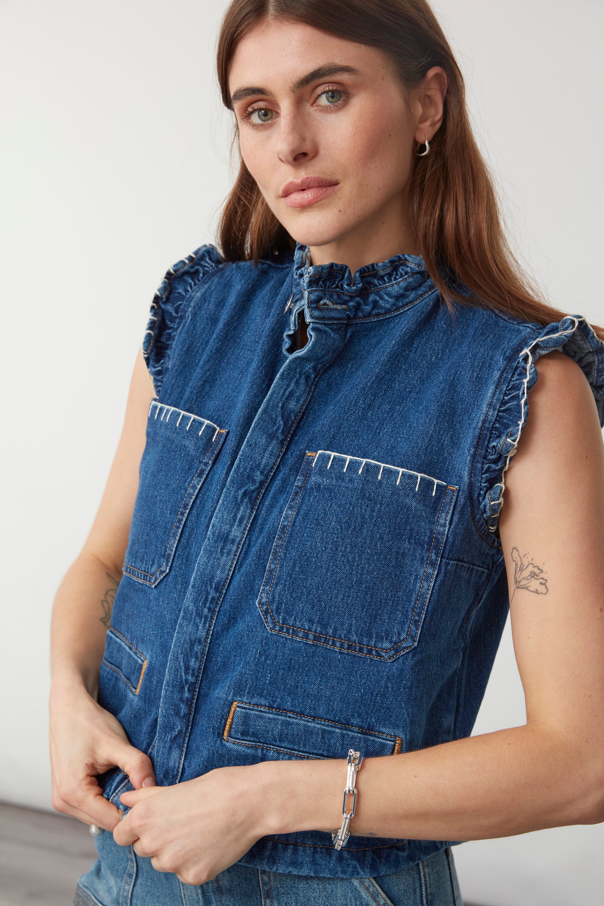Woman wearing a blue denim waistcoat/ vest with our signature white stitch detailing, two large patch pockets, and a frill collar against a white background