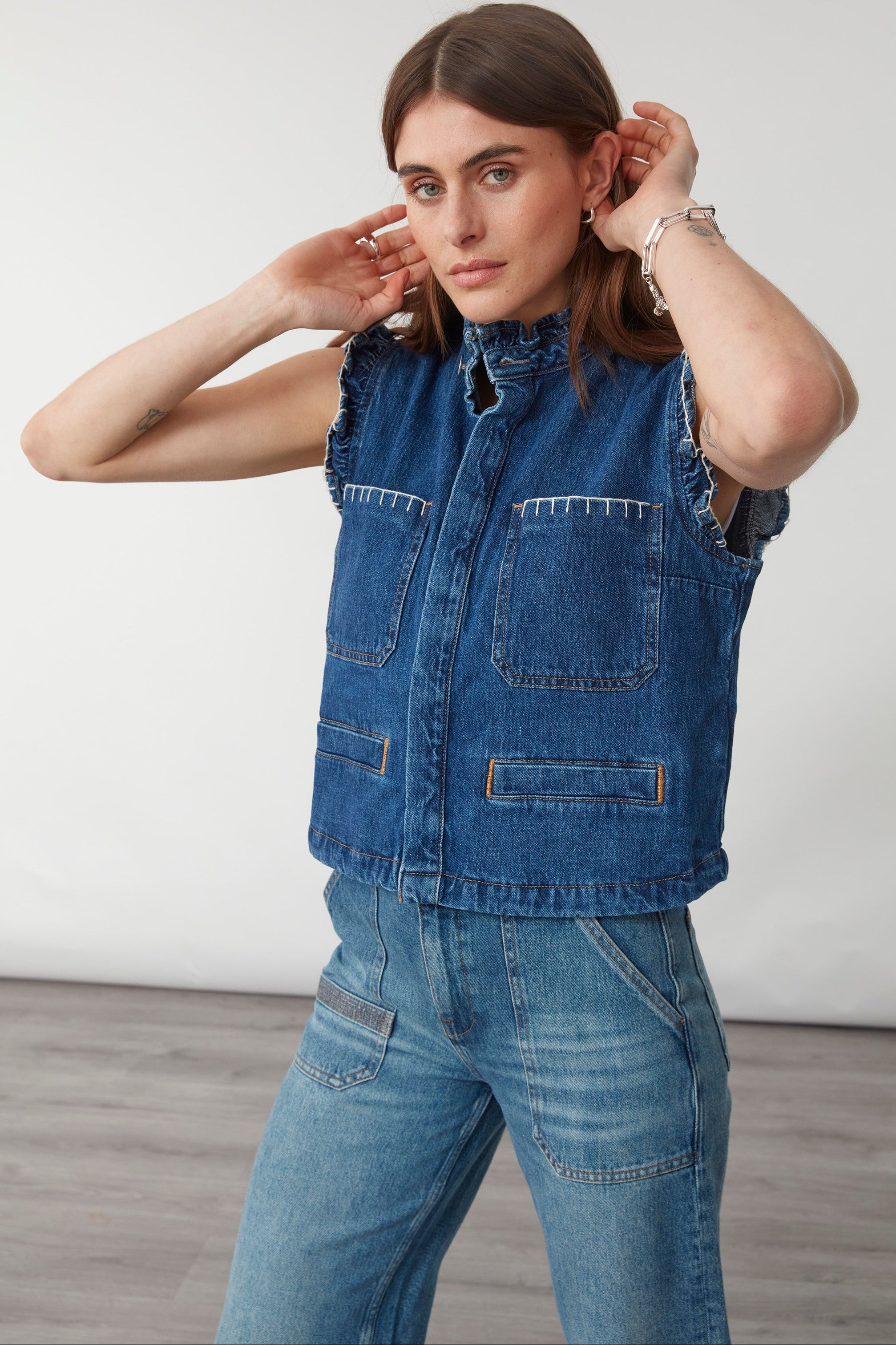 woman wearing a blue denim vest/ waistcoat with our signature white stitch detailing, two large patch pockets, and a frill collar with blue jeans against a white background
