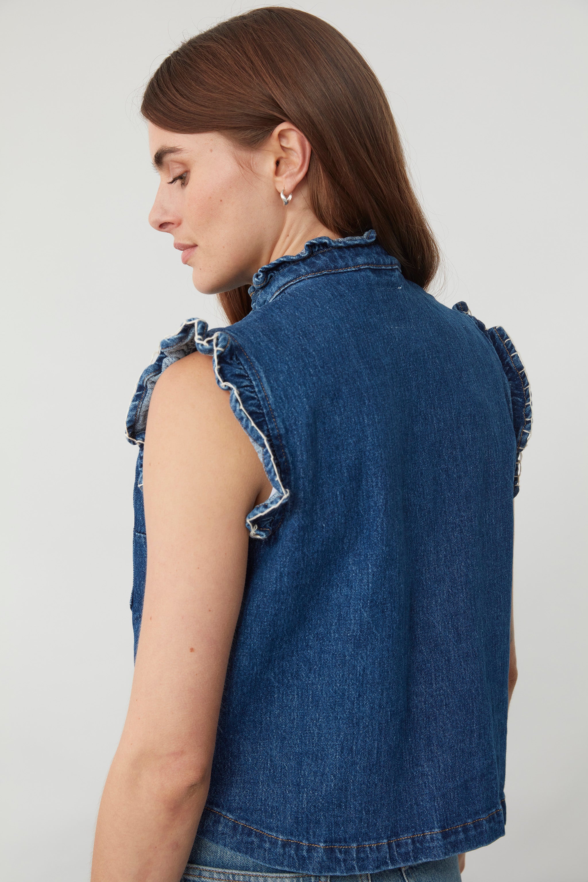 woman wearing a blue denim vest/ waistcoat with our signature white stitch detailing, two large patch pockets, and a frill collar against a white background