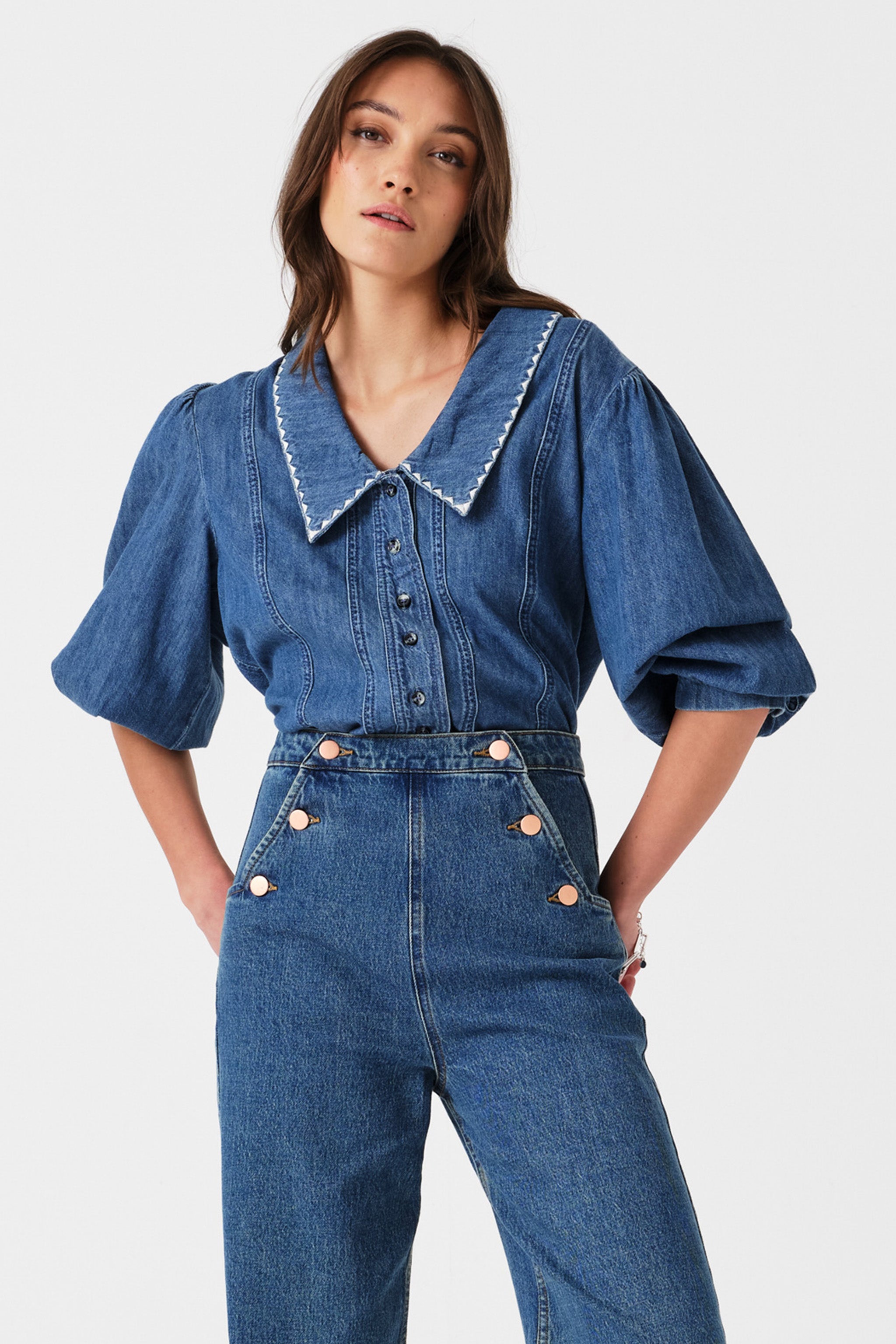 Woman wearing a blue denim button up top with high waisted jeans on a white background.