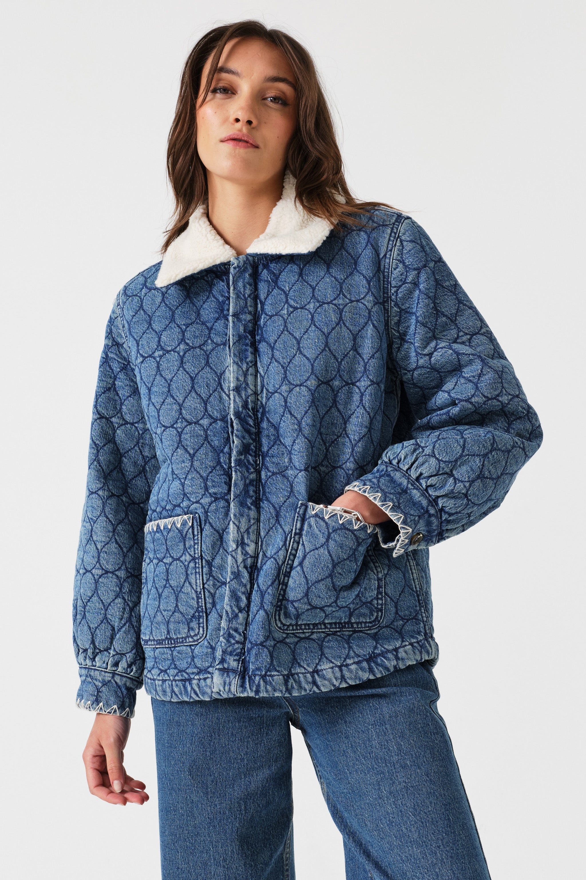 Woman wearing a blue quilted denim jacket with a removable white sherpa collar, large front pockets, and white embroidery details and blue jeans on a white background