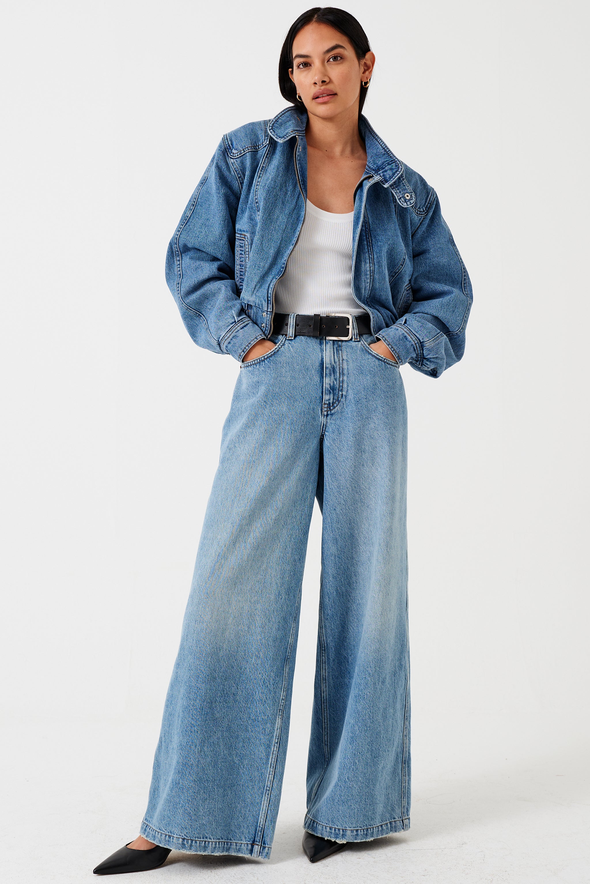 A woman wearing a high-rise, wide-leg, light wash blue denim jean, white shirt, and denim bomber jacket with a belt and black shoes against a white background