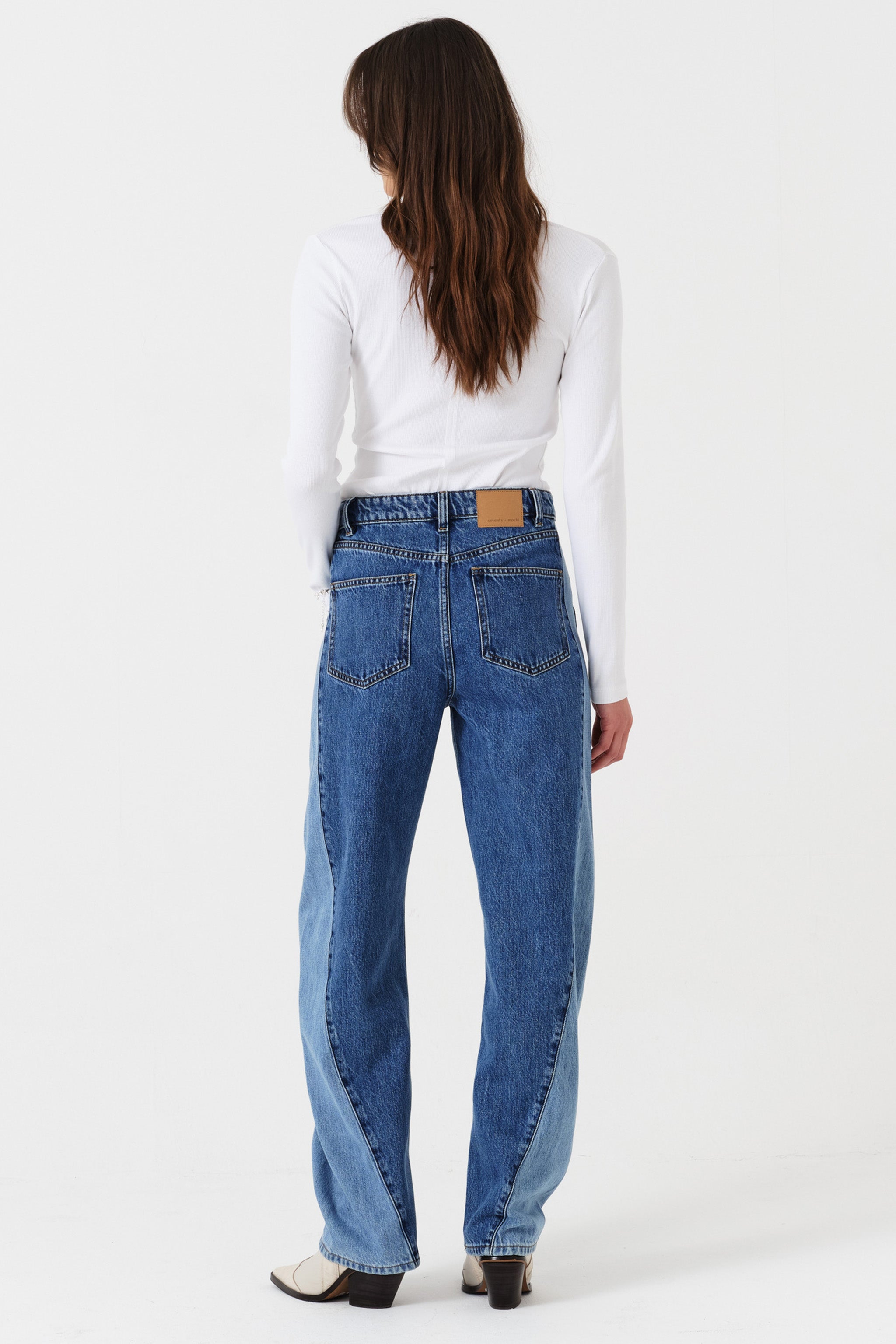 Woman wearing two toned blue barrel leg jeans and a white shirt on a white background back view