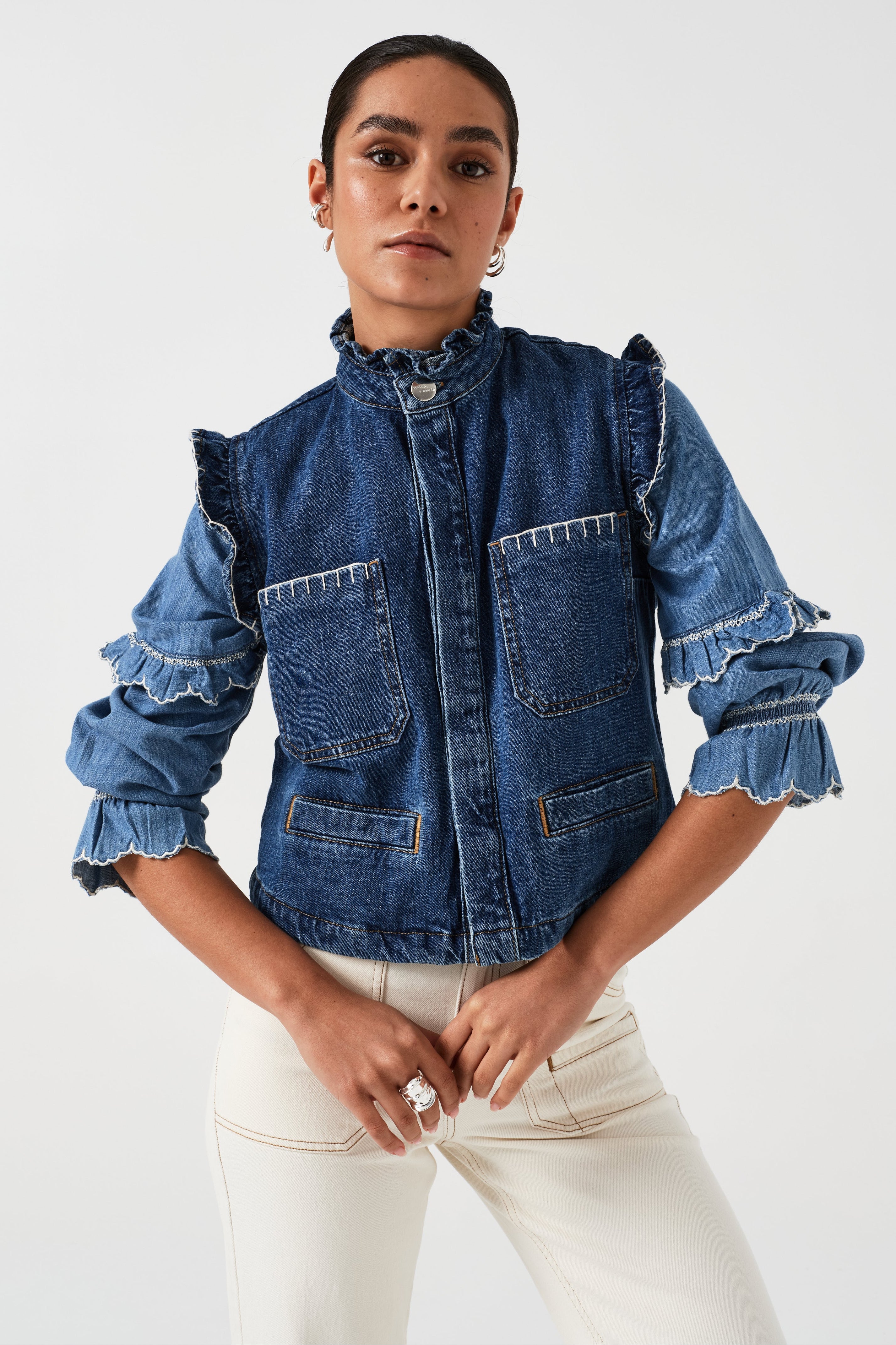 woman wearing a blue denim vest/ waistcoat with our signature white stitch detailing, two large patch pockets, and a frill collar with a blue shirt and white jeans against a white background