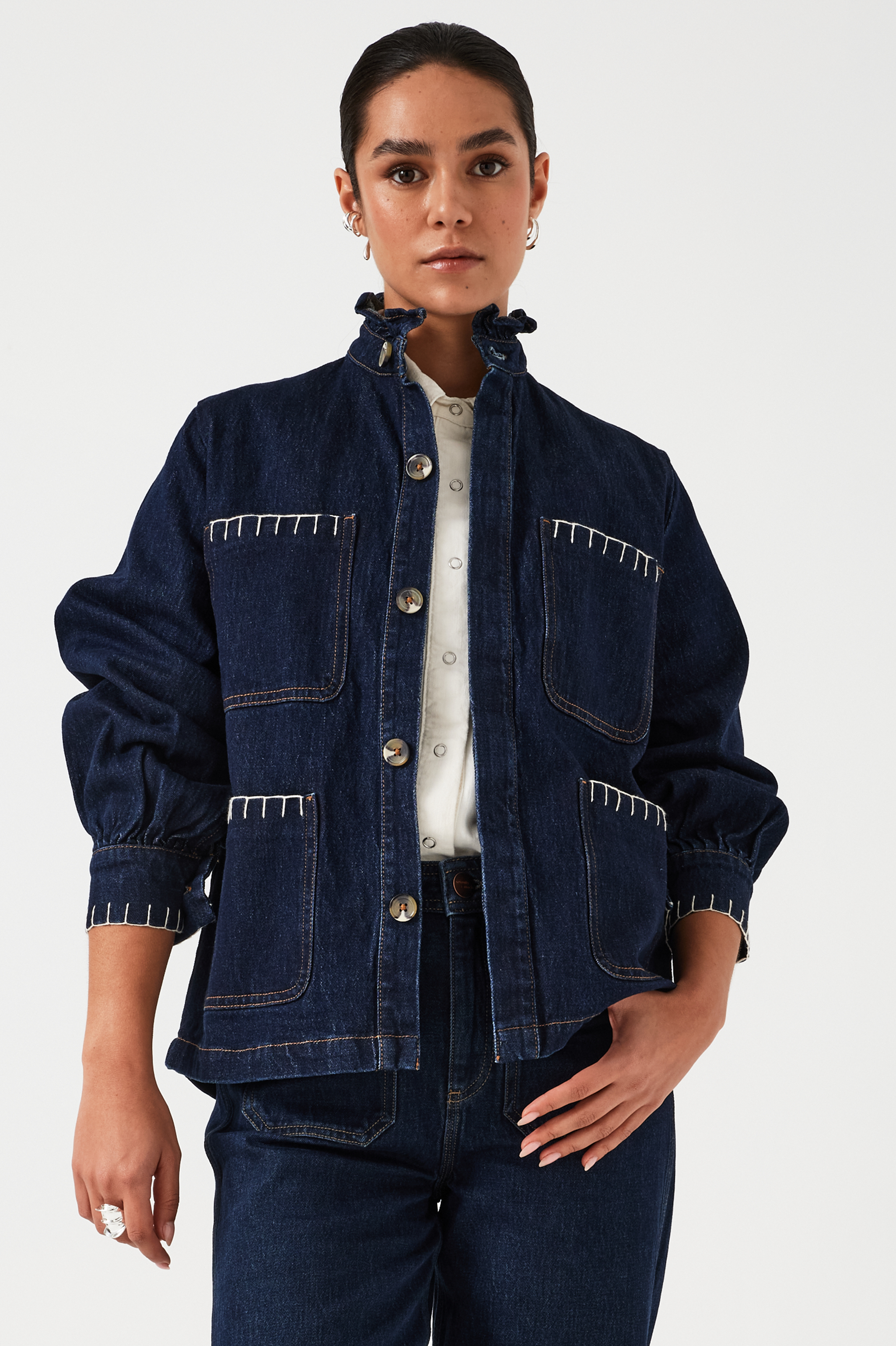 A woman wearing a dark blue vintage style jacket with a frill collar and our signature white blanket stitching on four large patch pockets, paired with a dark blue jeans and a white top against a white background
