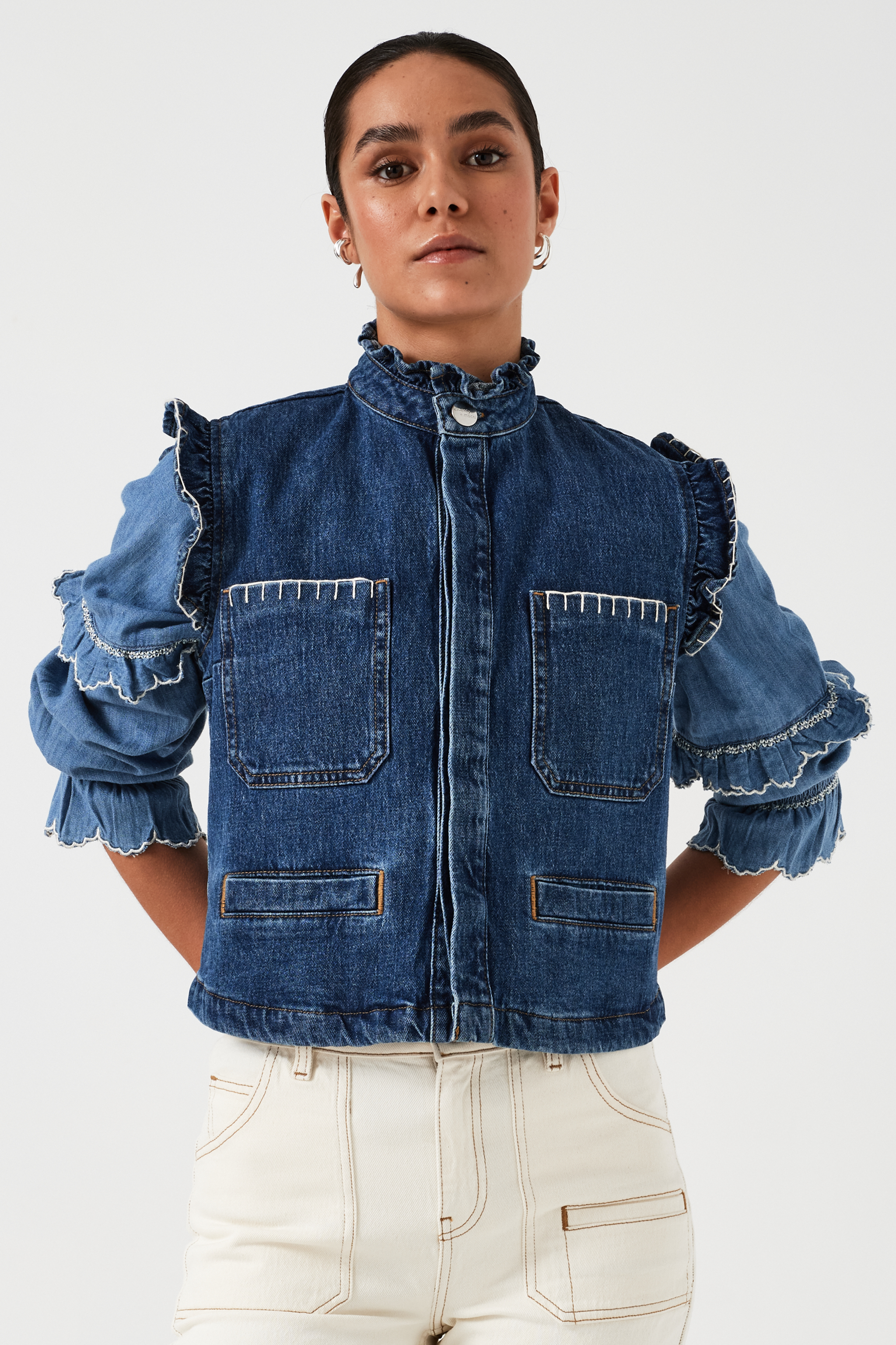 woman wearing a blue denim vest/ waistcoat with our signature white stitch detailing, two large patch pockets, and a frill collar with a blue shirt and white jeans against a white background